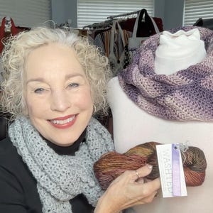 May include: A woman is holding a skein of yarn with a purple, brown, and gold color gradient. The yarn is labeled "Washed". The woman is wearing a gray crocheted scarf and a black shirt. A crocheted cowl in shades of purple and gray is displayed on a white mannequin.