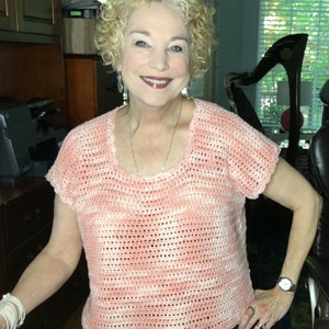 May include: A close-up of a woman wearing a pink and white crocheted top with short sleeves. The top has a loose, relaxed fit and a scoop neckline.
