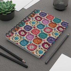 Pode incluir: Um caderno espiral com uma capa de croch&ecirc; colorida. A capa apresenta uma variedade de cores, incluindo vermelho, laranja, amarelo, verde, azul, roxo e rosa. O caderno est&aacute; sobre uma superf&iacute;cie cinza com dois l&aacute;pis pretos e uma folha de papel branca.