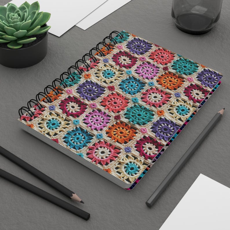 Pode incluir: Um caderno espiral com uma capa de croch&ecirc; colorida. A capa apresenta uma variedade de cores, incluindo vermelho, laranja, amarelo, verde, azul, roxo e rosa. O caderno est&aacute; sobre uma superf&iacute;cie cinza com dois l&aacute;pis pretos e uma folha de papel branca.