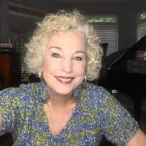 May include: A woman with curly blonde hair is wearing a blue, green, and white patterned shirt. She is smiling and looking at the camera. A black grand piano is in the background.