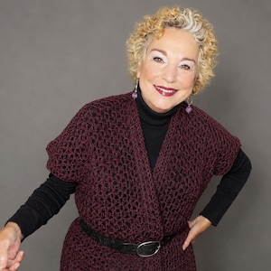 May include: A dark burgundy open-front vest featuring a loose, crocheted knit pattern with short sleeves. The garment is layered over a black long-sleeved mock neck shirt and is cinched at the waist with a black leather belt featuring a silver buckle.