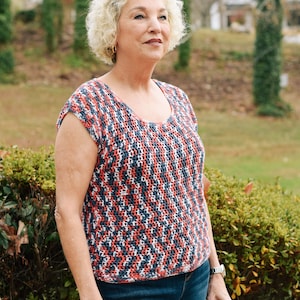May include: A woman with short, curly, blonde hair is wearing a red, white and blue crocheted top and blue jeans. She is standing in front of a green bush.