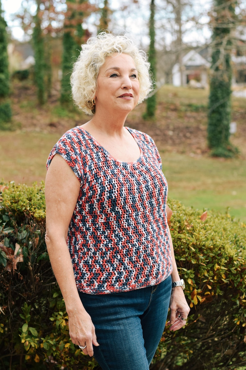 May include: A woman with short, curly, blonde hair is wearing a red, white and blue crocheted top and blue jeans. She is standing in front of a green bush.