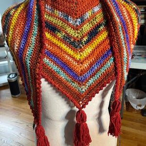 Peut inclure: Un châle crocheté à la main, coloré, avec un col montant et des glands. Le châle présente un mélange vibrant de fils orange, jaunes, bleus et violets. Les glands sont d'un rouge profond. Le châle est présenté sur un mannequin blanc.