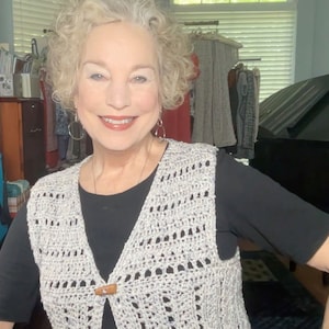 Puede incluir: Una mujer lleva un chaleco de ganchillo blanco con cierre de botón marrón sobre una camisa negra.