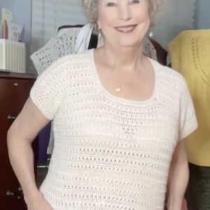May include: Cream-colored, short-sleeved crochet top with a scoop neck. The top features a textured stitch pattern and is paired with blue jeans. The overall style is casual and handmade.