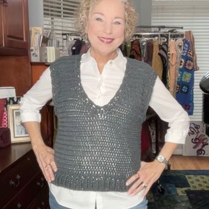 May include: A gray crocheted vest with a V-neckline. The vest is worn over a white button-down shirt and blue jeans.