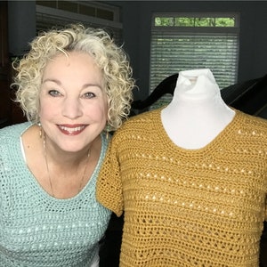May include: A woman wearing a light blue crochet top stands next to a crocheted top in a mustard yellow color. The top is on a white dress form.