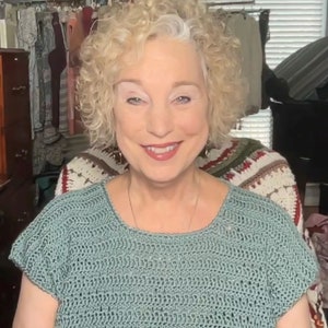 May include: A light teal crochet top with a round neckline and short sleeves. The top is made of a textured yarn and appears to be handmade. The woman is smiling and wearing a necklace.