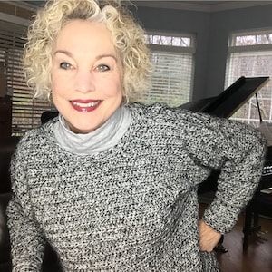 May include: A woman with curly blonde hair is wearing a black and white knit sweater. She is smiling and looking at the camera. She is standing in front of a grand piano.
