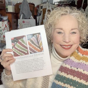 May include: A woman holds a printed pattern for a "Christmastime Coverlet" crochet project. The pattern shows two different color combinations for the crochet blanket. The woman is wearing a white turtleneck sweater and a colorful crocheted scarf.