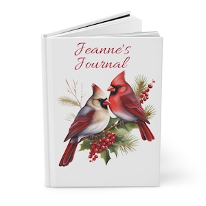 Pode incluir: Um diário branco com o texto "Jeanne's Journal" em escrita vermelha. A capa apresenta uma ilustração em aquarela de dois cardeais empoleirados em um galho com bagas vermelhas e folhas verdes. O diário está aberto.