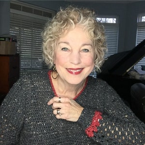 Może przedstawiać: Kobieta o kręconych blond włosach ma na sobie szary sweter z czerwonymi ozdobami zrobionymi na szydełku. Uśmiecha się i trzyma rękę na piersi. Ma na sobie złote kolczyki koła i kilka pierścionków.