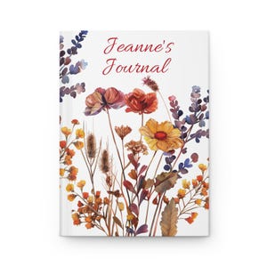 Pode incluir: Um diário branco com um design floral em aquarela. A capa apresenta uma variedade de flores em tons de laranja, vermelho e amarelo, com folhagem roxa e verde. O texto "Jeanne's Journal" está escrito em vermelho.