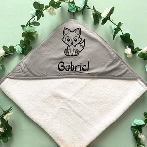 May include: A grey hooded towel with a white fox design and the name "Gabriel" embroidered on it. The towel is on a green background with a sign that reads "BOUILLE D'AMOUR COM".
