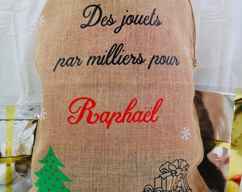May include: A brown burlap sack with a drawstring closure. The sack has a Christmas tree and a stack of presents printed on it. The text "Des jouets par milliers pour Raphael" is printed on the sack.