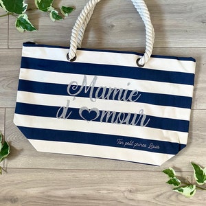 Puede incluir: Una bolsa de tela de rayas blancas y azules con asa de cuerda. La bolsa tiene el texto "Mamie d'amour" en letras plateadas con purpurina y el texto "Ton petit prince Louis" en letras plateadas con purpurina m&aacute;s peque&ntilde;as.