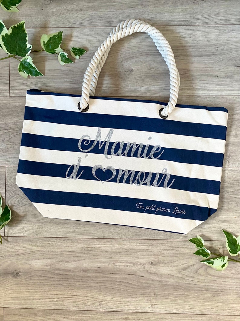 Puede incluir: Una bolsa de tela de rayas blancas y azules con asa de cuerda. La bolsa tiene el texto "Mamie d'amour" en letras plateadas con purpurina y el texto "Ton petit prince Louis" en letras plateadas con purpurina m&aacute;s peque&ntilde;as.