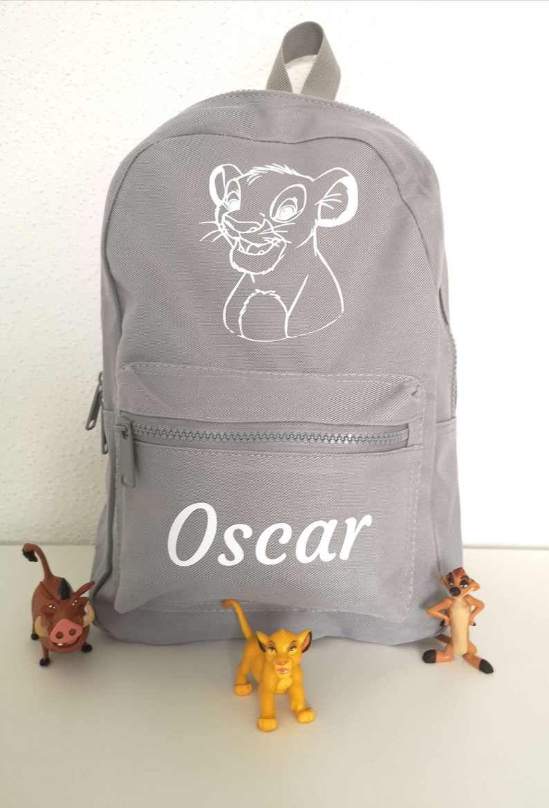 K&ouml;nnte beinhalten: Ein hellgrauer Rucksack mit einer wei&szlig;en Linienzeichnung eines L&ouml;wenjungen im oberen Bereich und dem Namen Oscar in wei&szlig;em Druck auf der vorderen Rei&szlig;verschlusstasche. Der Rucksack hat einen Tragegriff und passende graue Rei&szlig;verschl&uuml;sse.