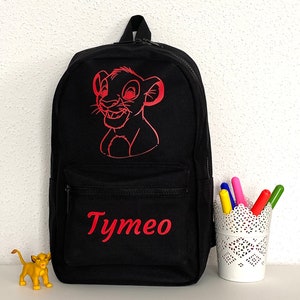 K&ouml;nnte beinhalten: Ein schwarzer Rucksack mit einer roten Strichzeichnung eines L&ouml;wenjungen im oberen Bereich und dem Namen Tymeo in roter Schrift auf der vorderen Rei&szlig;verschlusstasche. Eine kleine gelbe L&ouml;wenbaby-Figur und ein wei&szlig;er Stiftehalter stehen daneben.