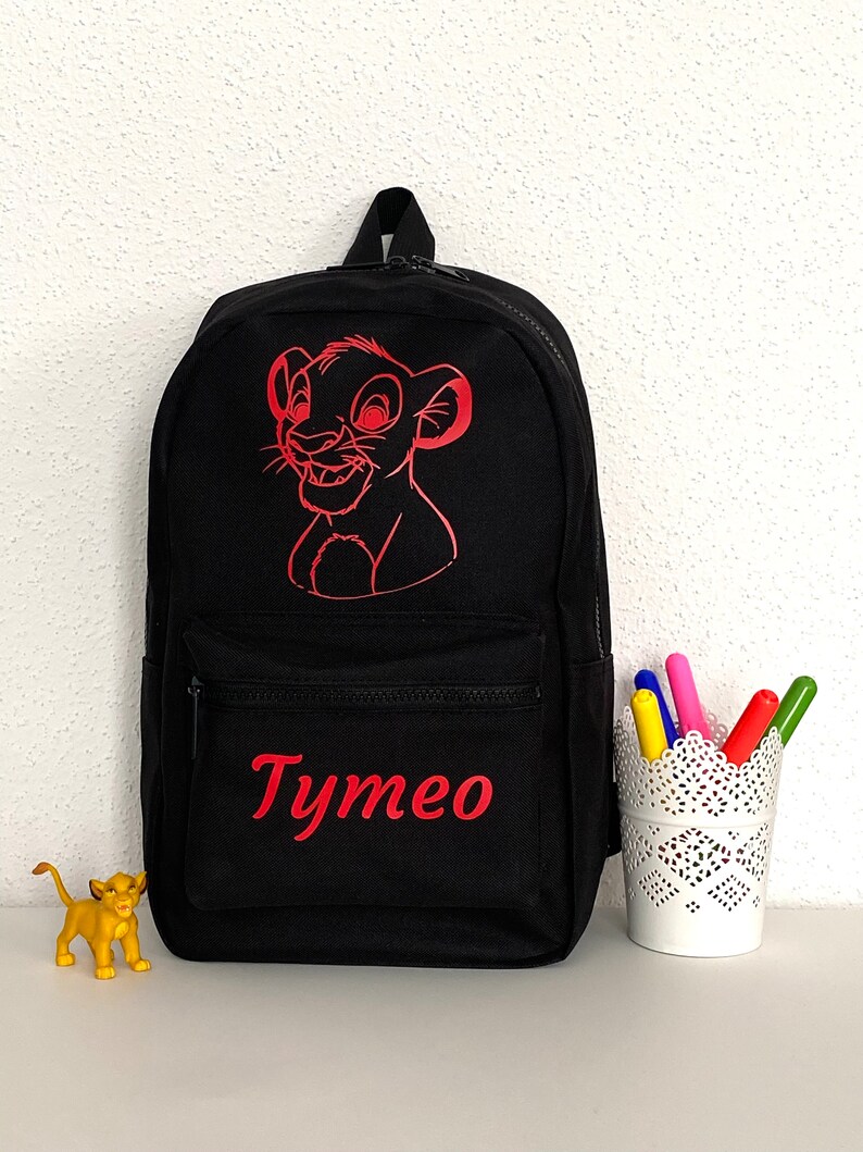 K&ouml;nnte beinhalten: Ein schwarzer Rucksack mit einer roten Strichzeichnung eines L&ouml;wenjungen im oberen Bereich und dem Namen Tymeo in roter Schrift auf der vorderen Rei&szlig;verschlusstasche. Eine kleine gelbe L&ouml;wenbaby-Figur und ein wei&szlig;er Stiftehalter stehen daneben.