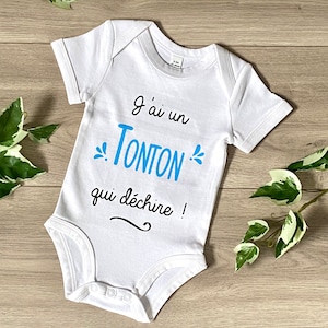 Könnte beinhalten: Weißer Baby Strampler mit blauem Text, der "J'ai un Tonton qui déchire!" lautet, was übersetzt "Ich habe einen tollen Onkel!" bedeutet.
