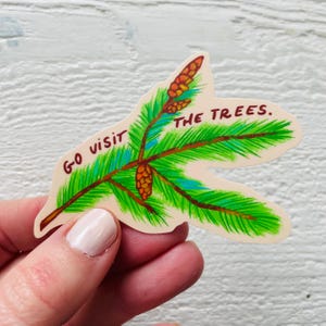 Peut inclure: Un autocollant représentant une branche de pin stylisée avec des aiguilles vertes, des branches marron et des cônes orange. Le texte "GO VISIT THE TREES" est écrit en bordeaux. L'autocollant est tenu par une personne.