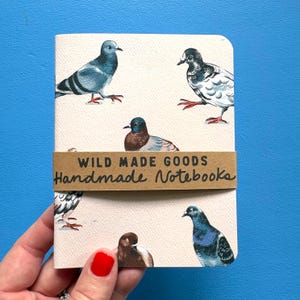 May include: A cream-coloured notebook featuring a repeating pattern of various pigeons in shades of blue, grey, and brown. A brown paper band wraps around the centre with the black printed text "WILD MADE GOODS" and "Handmade Notebooks".