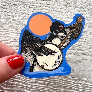 May include: A sticker depicting a cartoon loon playing a banjo. The loon is black and white with a red eye, set against an orange sun and blue water. The sticker is held by a hand with red nail polish.