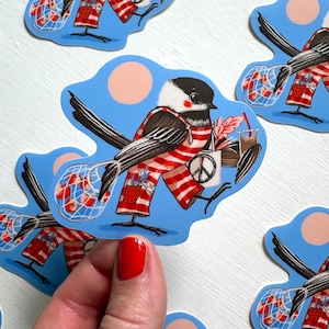 May include: A die-cut sticker featuring a black and white chickadee wearing red and white striped overalls. The bird carries a mesh bag filled with red apples, a white tote bag with a black peace sign, and a brown beverage in a clear cup.