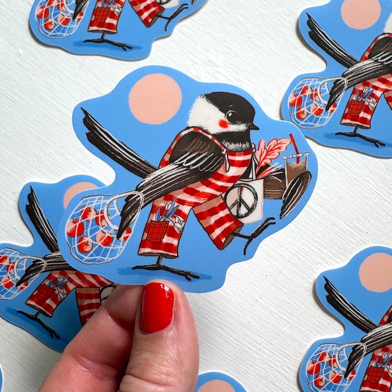 May include: A die-cut sticker featuring a black and white chickadee wearing red and white striped overalls. The bird carries a mesh bag filled with red apples, a white tote bag with a black peace sign, and a brown beverage in a clear cup.