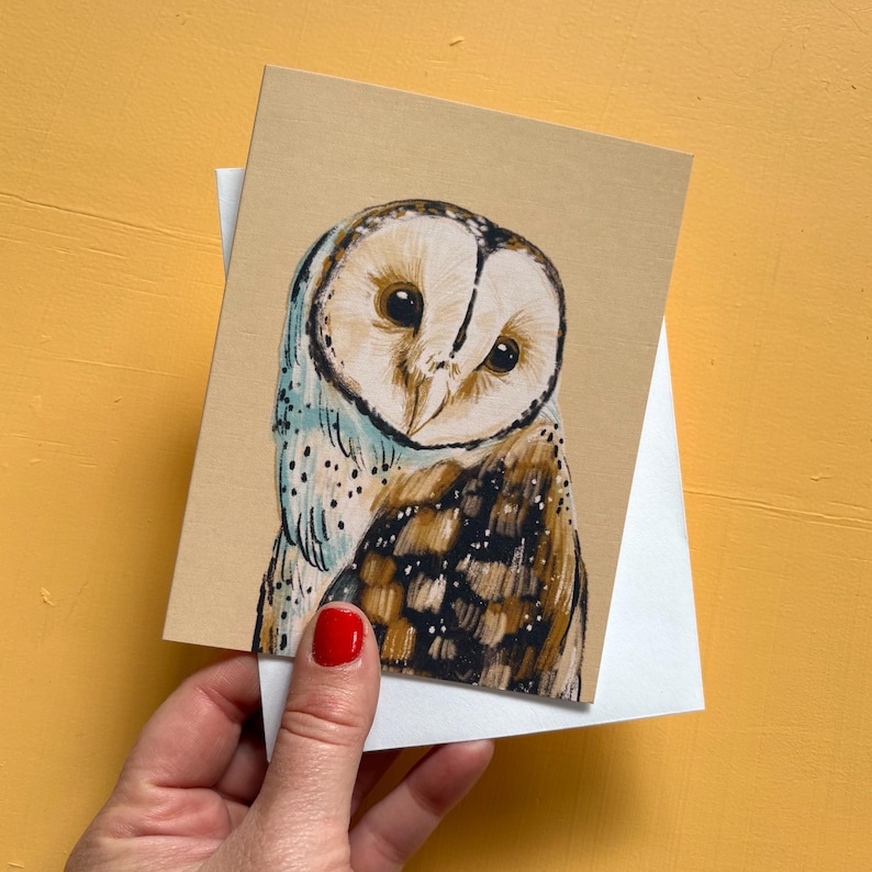 May include: A rectangular greeting card featuring a detailed illustration of a barn owl with a white heart-shaped face, dark eyes, and brown and white speckled feathers on a light tan background, resting on a plain white envelope.