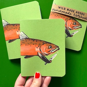 Peut inclure: Trois carnets vert clair illustrés d'une truite orange tenant un petit couteau argenté. L'un des carnets porte une bande en papier brun avec le texte WILD MADE GOODS Handmade Notebook imprimé en noir sur la couverture avant.