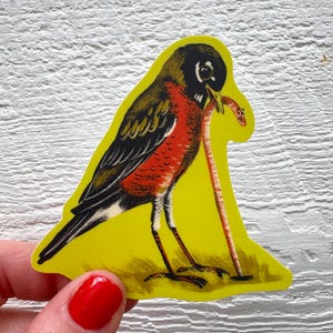 May include: A yellow sticker featuring a stylized illustration of a robin bird with a worm in its beak. The bird has a black head, brown wings, and a red breast. The worm is a light orange color.
