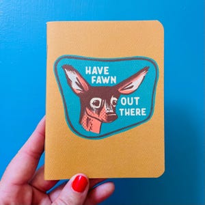 May include: A mustard-coloured notebook with rounded corners. The cover features a teal patch with a deer illustration and the words "HAVE FAWN OUT THERE" in white. The notebook is held by a hand with red nail polish.
