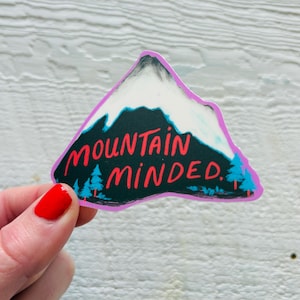 May include: A mountain-shaped sticker featuring a black and white mountain design, the red text "MOUNTAIN MINDED", and blue trees. The sticker has a purple border and is held by a hand with red nail polish.
