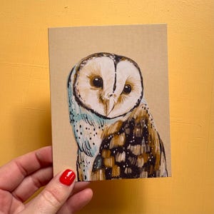 May include: A rectangular greeting card featuring a detailed illustration of a barn owl. The owl has a heart-shaped white face, dark eyes, and brown, black, and white feathers. The artwork is set against a plain, light tan background.