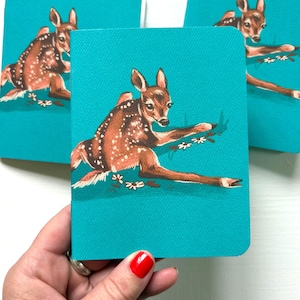 May include: Three teal cards with a vintage illustration of a fawn. The fawn is brown with white spots, lying on grass with small white flowers. The cards are held in a hand. The fawn has large ears and is set against a teal background.