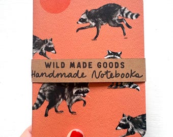 May include: A rectangular notebook with a coral orange cover featuring a repeating pattern of black and white raccoons and a solid orange circle. A brown paper band wraps around the center with the text WILD MADE GOODS Handmade Notebooks.