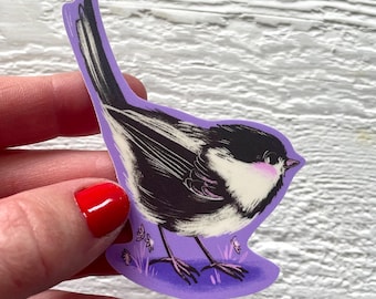May include: A sticker featuring a detailed illustration of a small bird with black and white feathers, a pink cheek, and a black head. The bird is set against a purple background with small flowers. The sticker is held by a person.
