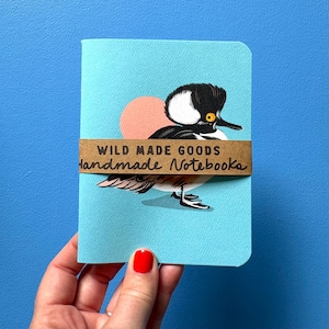 May include: A light blue notebook featuring a graphic illustration of a hooded merganser duck with a black and white crest and a yellow eye. A brown paper band wraps around the centre with the text WILD MADE GOODS Handmade Notebooks.