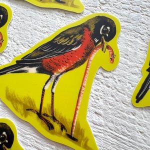 May include: Yellow die-cut sticker featuring a stylized bird with a red breast and a worm in its beak. The bird has black and brown wings and is set against a yellow background. The sticker is likely part of a set.