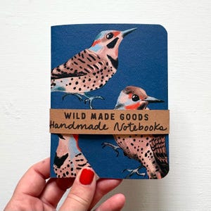 May include: A small notebook with a dark blue cover featuring a pattern of spotted birds with red and black markings. A brown paper band wraps around the centre with the text WILD MADE GOODS Handmade Notebooks printed in black ink.