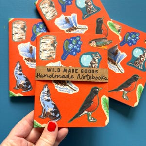May include: Handmade notebooks with an orange background and nature-themed illustrations. The notebooks feature images of a howling wolf, a robin, a fish, and a birch log. The text on the notebooks reads "Wild Made Goods Handmade Notebooks."