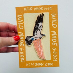 May include: Embroidered patch of a bird in flight, with brown, white, and peach hues. The patch is set on a cream background, within a mustard-yellow frame. The frame has the text "WILD MADE GOODS".