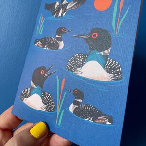 Pode incluir: Um fundo azul com um padr&atilde;o repetido de p&aacute;ssaros mergulhadores pretos e brancos nadando na &aacute;gua. Os p&aacute;ssaros t&ecirc;m olhos vermelhos e est&atilde;o virados em dire&ccedil;&otilde;es diferentes. Tamb&eacute;m h&aacute; juncos verdes e um sol vermelho no fundo.