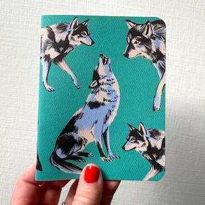 Peut inclure: Un carnet de notes turquoise avec un motif répété de loups noirs, blancs et bleu clair. Un loup hurle, les autres sont dans différentes poses. Le carnet est tenu par une main avec du vernis à ongles rouge.