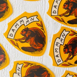 May include: Four yellow and white shield-shaped stickers with a cartoon beaver holding a banner that reads "DAMM MN". The beaver is brown and orange with black eyes and a black nose. The background is a white wooden surface.