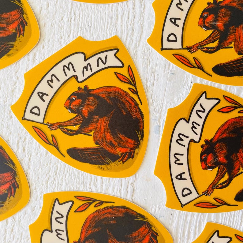May include: Four yellow and white shield-shaped stickers with a cartoon beaver holding a banner that reads "DAMM MN". The beaver is brown and orange with black eyes and a black nose. The background is a white wooden surface.